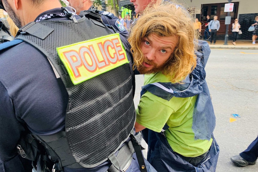 A man with long hair looks back at the camera as he's led away by police.