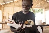 An Aboriginal man holding a carved wooden dog inside a workshed.