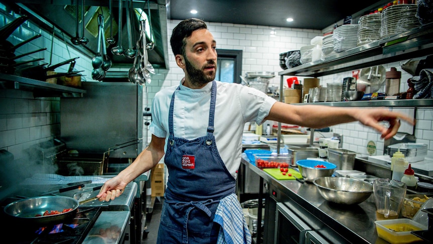 A chef fries mushrooms in a restaurant kitchen and waves with his other hand