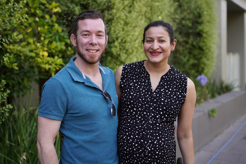 A man wearing a blue polo shirt with a woman wearing a spotty black dress in front of a hedge