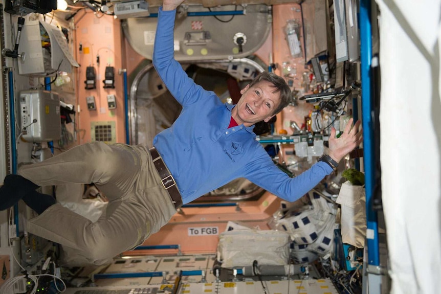 A smiling woman floats in an enclosed area of a space station. Her hands are in the air.