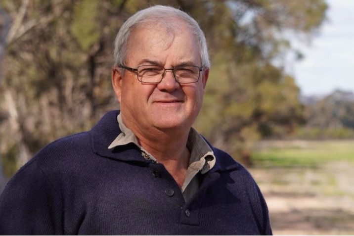 A bespectacled, middle-aged man with short grey hair stands in a country setting.