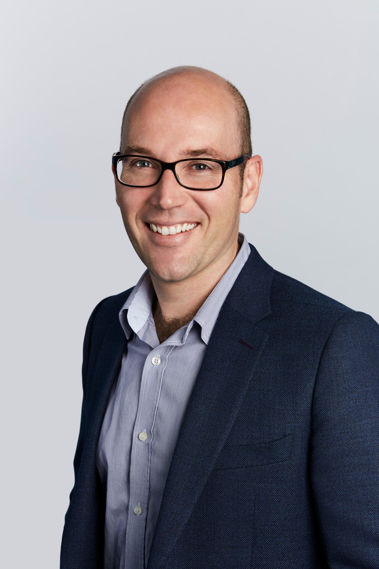 A balding man wearing glasses, an open-neck business shirt and suit jacket smiles for the camera.