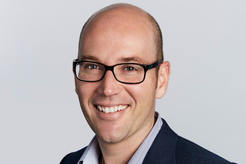 A balding man wearing glasses, an open-neck business shirt and suit jacket smiles for the camera.