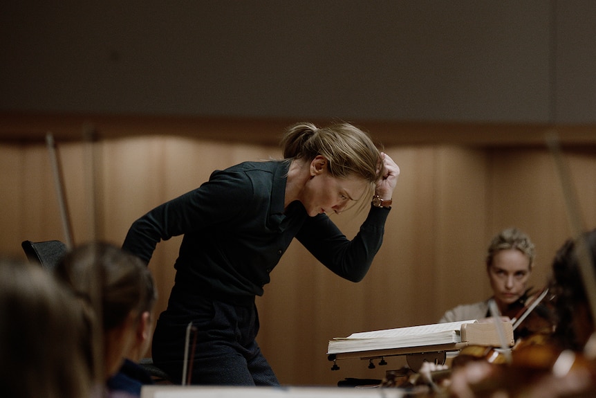 A brown-haired, 50-something woman leans forward, hand in a fist, with a look of concentration, in front of a group of musicians