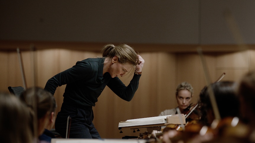 A brown-haired, 50-something woman leans forward, hand in a fist, with a look of concentration, in front of a group of musicians