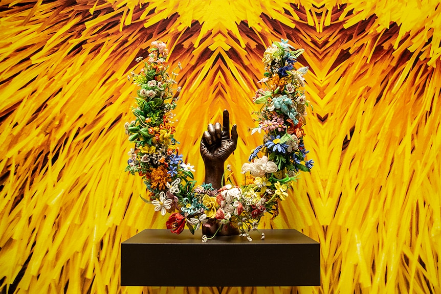 Colour photo of a bronze arm, U-shaped floral wreath in front of wall-sized photograph of vibrant yellow textures.