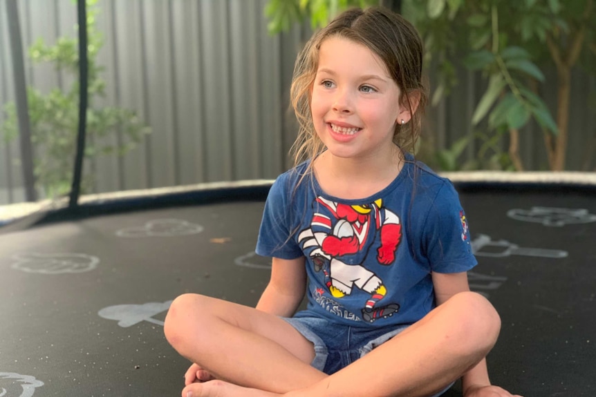 A young girl sits cross-legged and smiling on a trampoline.