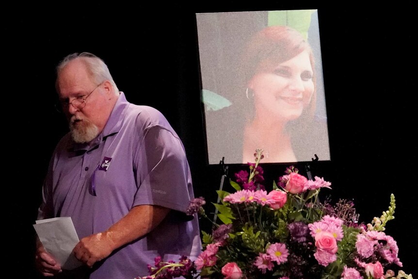 A man in a purple shirt walks between a bouquet of flowers and a large photo of his daughter.