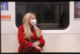 A woman in a red coat sitting on a train wearing a protective face mask.