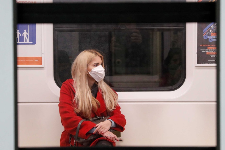 A woman in a red coat sitting on a train wearing a protective face mask.
