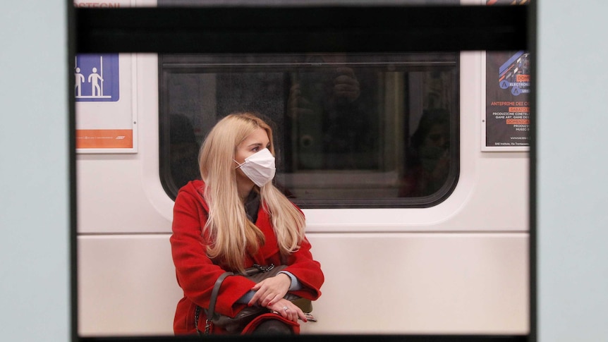 A woman in a red coat sitting on a train wearing a protective face mask.