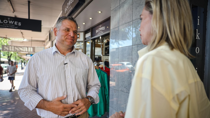 A man in a shirt talks to a woman outside a shop.