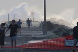 People walk past a 'Beach Closed' sign at the Southport Spit while large waves crash in the background.