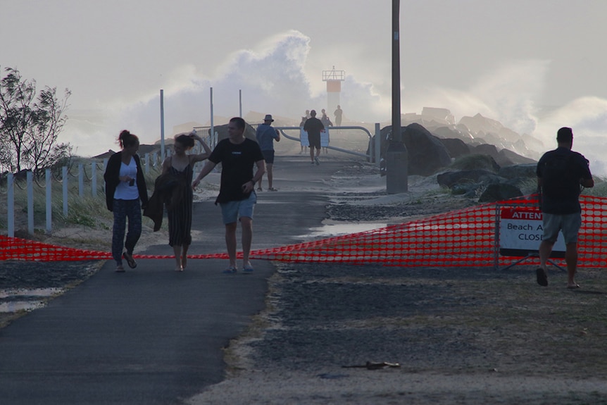 People walk past a 'Beach Closed' sign at the Southport Spit while large waves crash in the background.