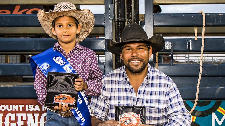 Little boy and older man, both Aboriginal and wearing cowboy clothes, hold up rodeo buckle prizes