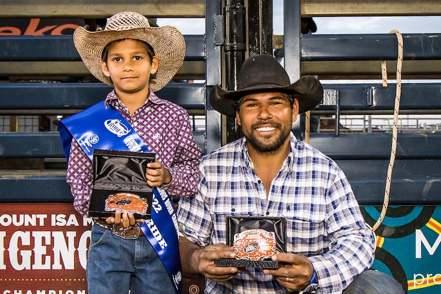 Little boy and older man, both Aboriginal and wearing cowboy clothes, hold up rodeo buckle prizes