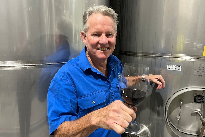 Anthony Murphy holds a glass of red wine while standing in front of some stainless steel wine tanks