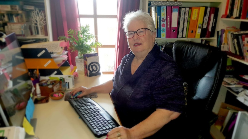 A woman with short white hair sits in a small office, surrounded by books, in front of a bright window.