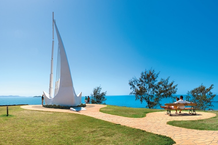 The 12-metre-tall Singing Ship overlooks the blue ocean Keppel Bay with people nearby on a park bench enjoying the view.