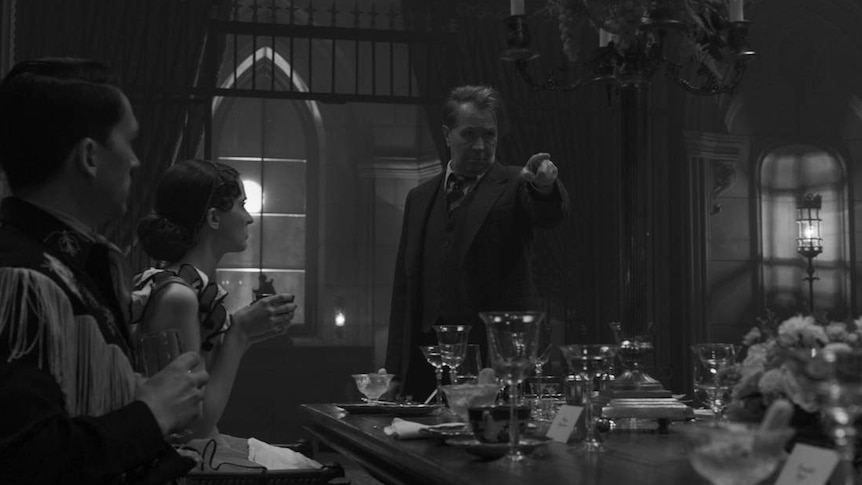 Black and white film still showing Gary Oldman in 30s suit standing at foot of dining table pointing to its unseen end.