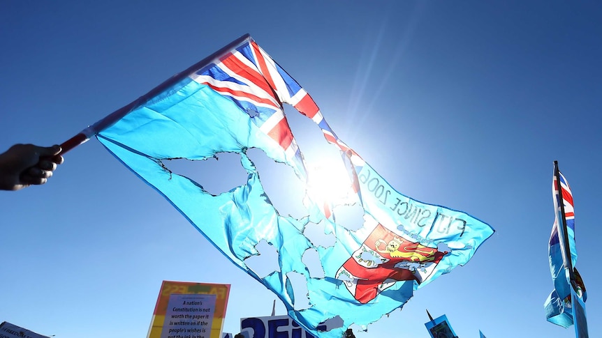 A Fijian flag that is burnt is held aloft into the sky