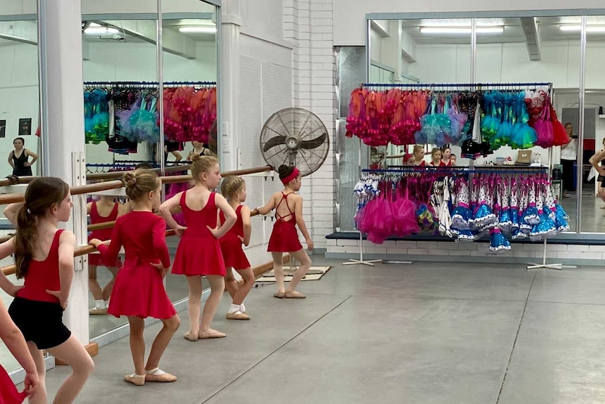 Young ballerinas in red and black leotards dance in a mirrored ballet studio with colourful costumes in the background.