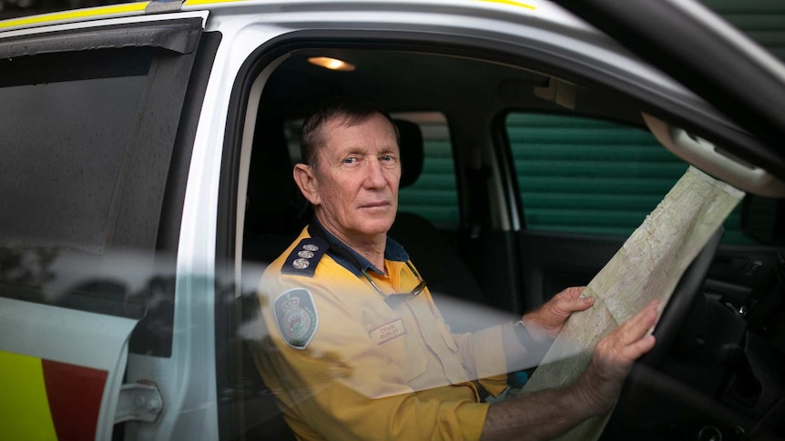 A middle-aged man in a firefighter's uniform sits in an emergency vehicle, holding up a map.