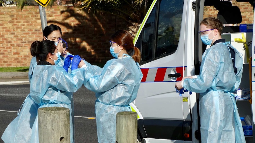 A group of women wearing blue PPE gear and wearing masks stand in the street.