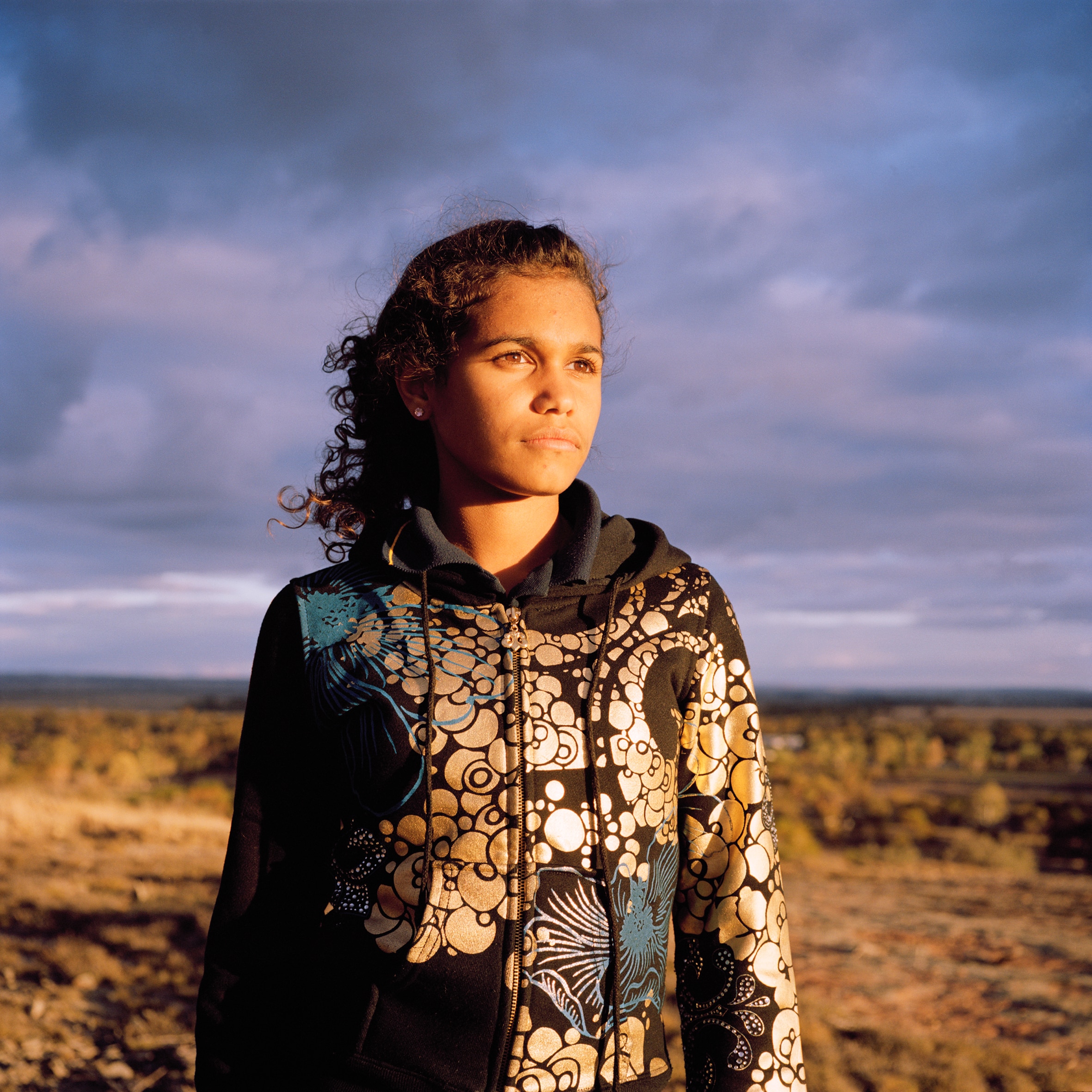 Young woman in hoodie stands outside in a scrubby landscape, with dark clouds behind her