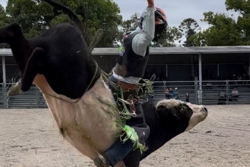 A teenage boy sits atop a bucking bull with one hand in the air and the bull is kicking its hind legs in the air. 