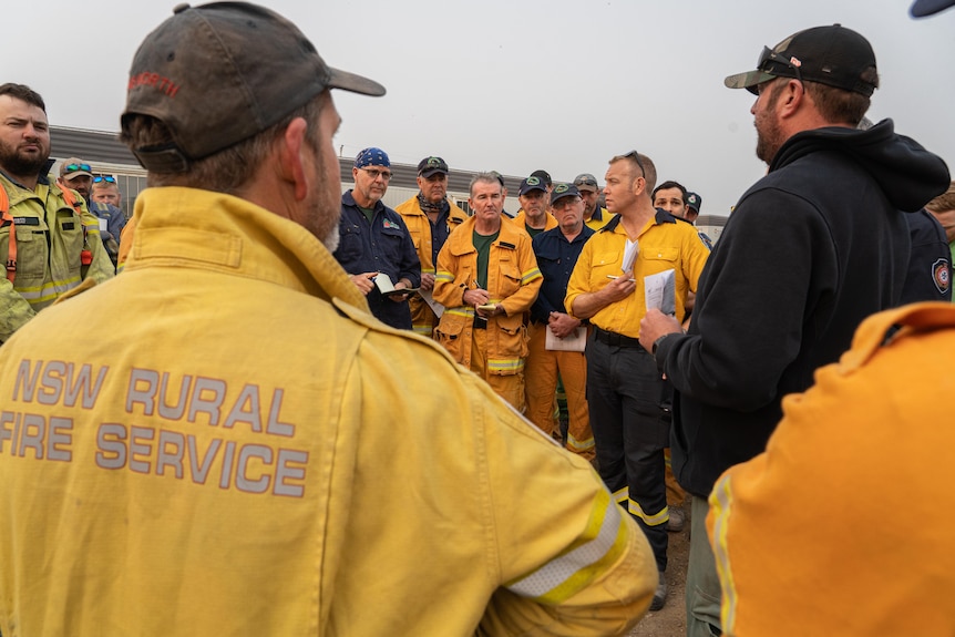 A group of firefighters wearing yellow uniforms gather for their morning briefing outside.