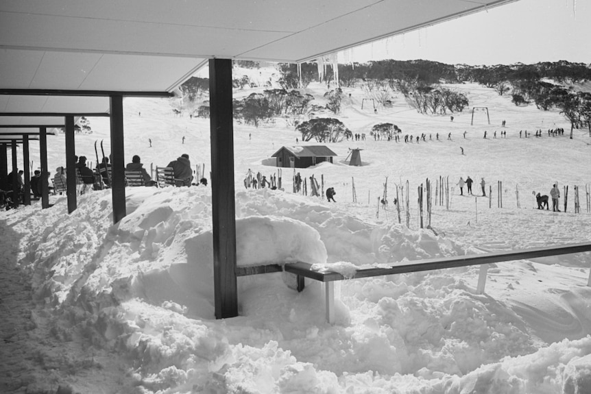 Chalet heaped with large piles of snow and with people skiing on the slopes beyond