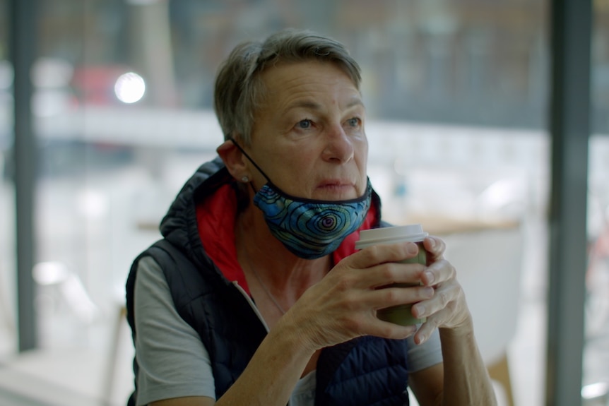 A woman sits, staring off into the distance, holding a cup of coffee
