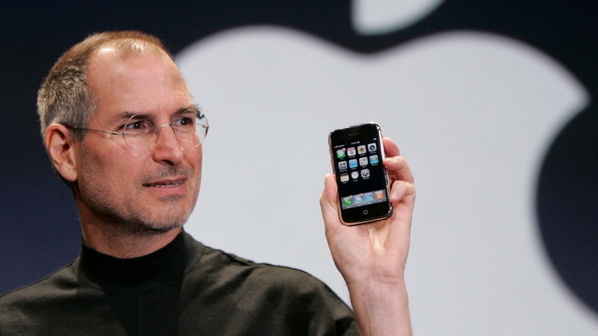 A man wearing glasses holds up an iPhone in his hand.