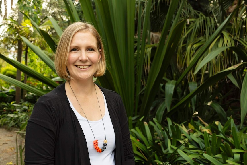 A smiling blonde woman, apparently in her 30s, standing in front of a large succulent.