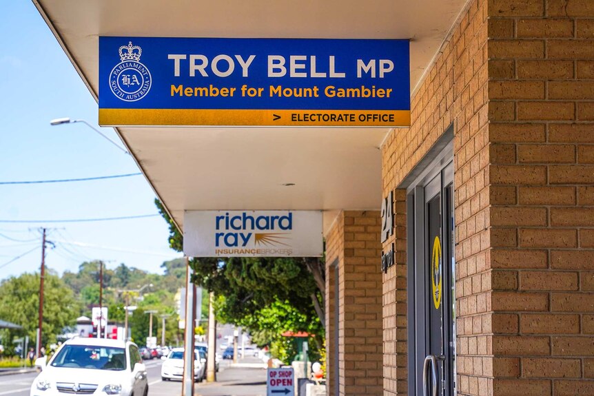 A sign hanging from a walkway next to a brick building reads 'Troy Bell MP, Member for Mount Gambier, electorate office'.