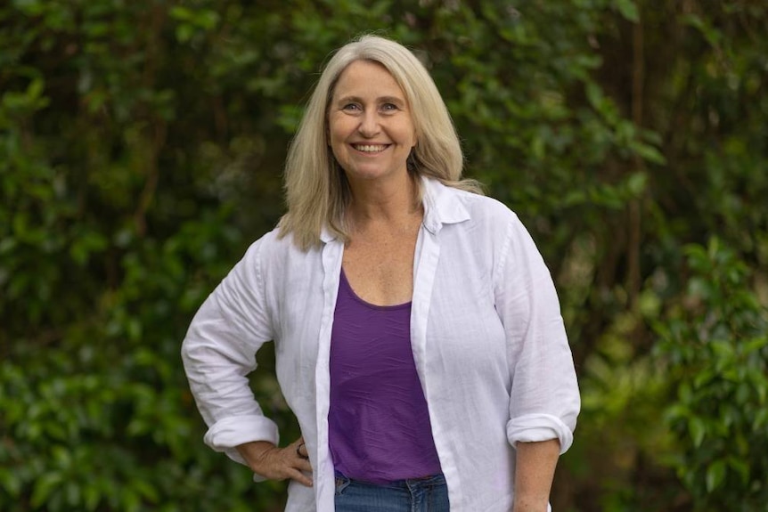 A smiling blonde lady, wearing a purple and white outfit with her hand on her hip 