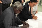 Tiwi Plantations Corp Kim Puruntatameri sits beside Mitsui MD Yasuhiro Yamano at a table and sign documents