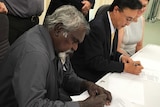 Tiwi Plantations Corp Kim Puruntatameri sits beside Mitsui MD Yasuhiro Yamano at a table and sign documents