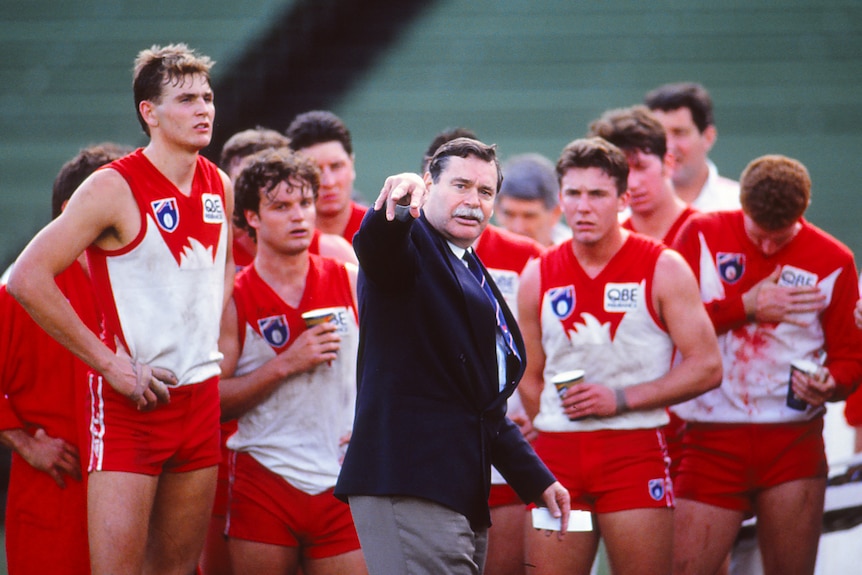 A Sydney Swans AFL coach points across the ground during a team talk with his players gathered behind him on the ground.