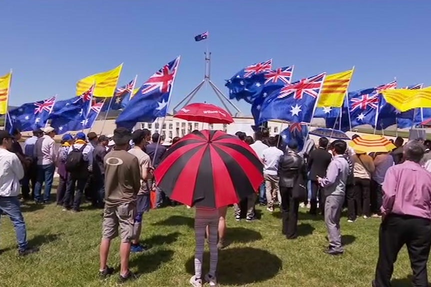 A group of people with Australian flags, yellow and red striped flags stand in front of Parliament House.