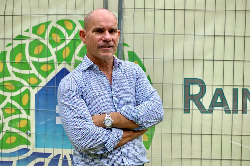 Man wearing blue checked short standing in front of temporary fencing with his arms crossed.