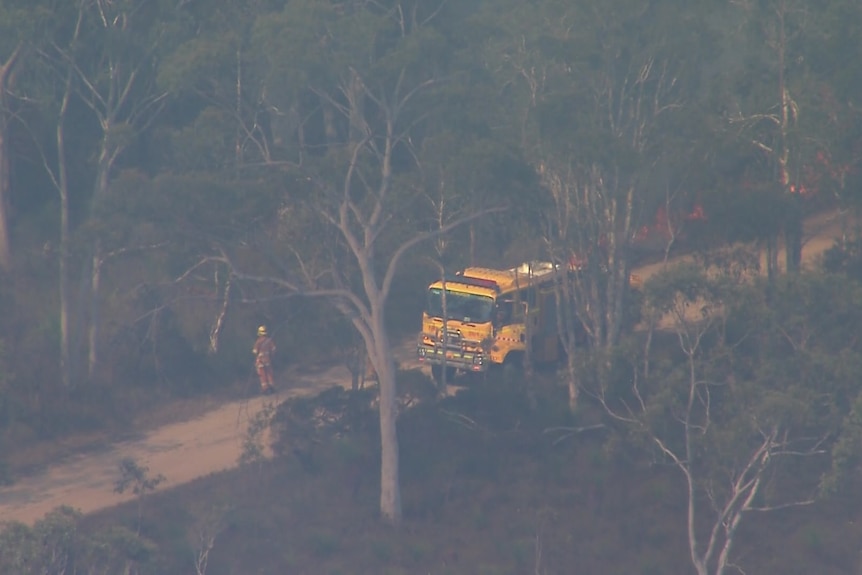 an aerial photo of a firefighter standing next to a yellow fire truck assessing a grass fire in thick bushland