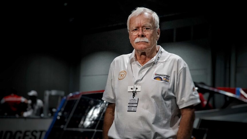 Man with white hair and white moustache stands in front of a solar car.