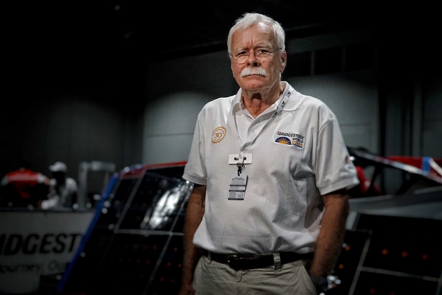 Man with white hair and white moustache stands in front of a solar car.