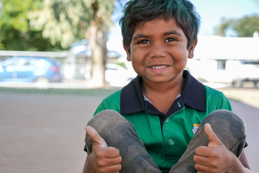 A young Indigenous boy sits holding his legs up, grinning and giving both thumbs up.