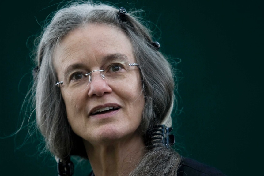 A bespectacled Sharon Olds looks into the camera in front of a dark green background.