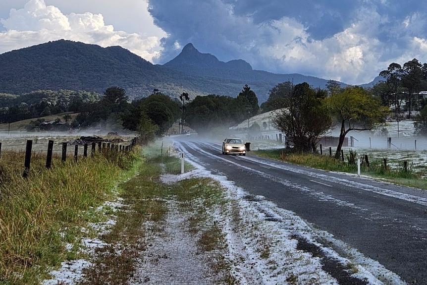 A car drives along a road scattered with hail. Clouds loom over mountains in the background.