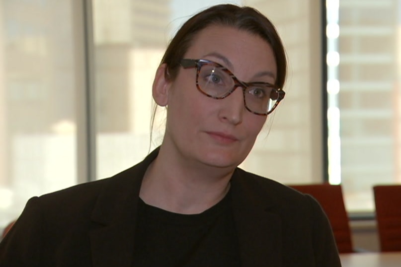 A woman with dark hair wears glasses and a black suit and looks sternly away from the camera